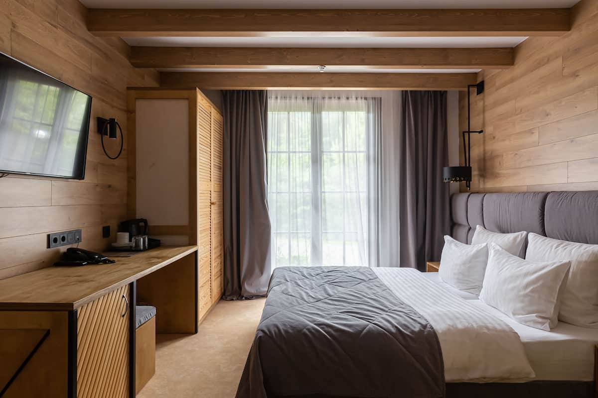 A modern hotel room with a wooden interior, a bed with white bedding, a desk with a coffee maker, and a television mounted on the wall.