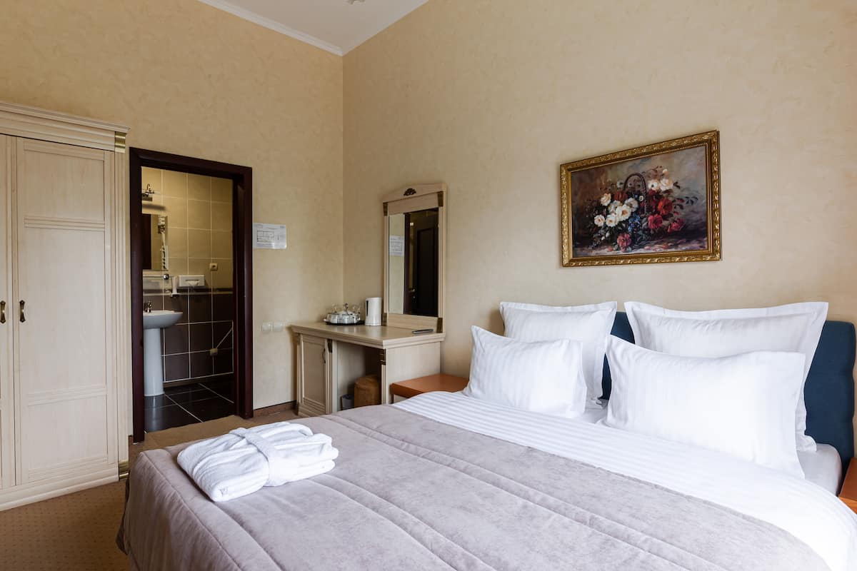 A hotel room with a bed, a desk with a mirror, a painting, and a wardrobe.