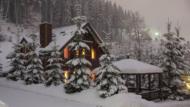 Une cabane en bois, dont le toit est recouvert de neige, entourée d’arbres et illuminée par une lumière chaleureuse.