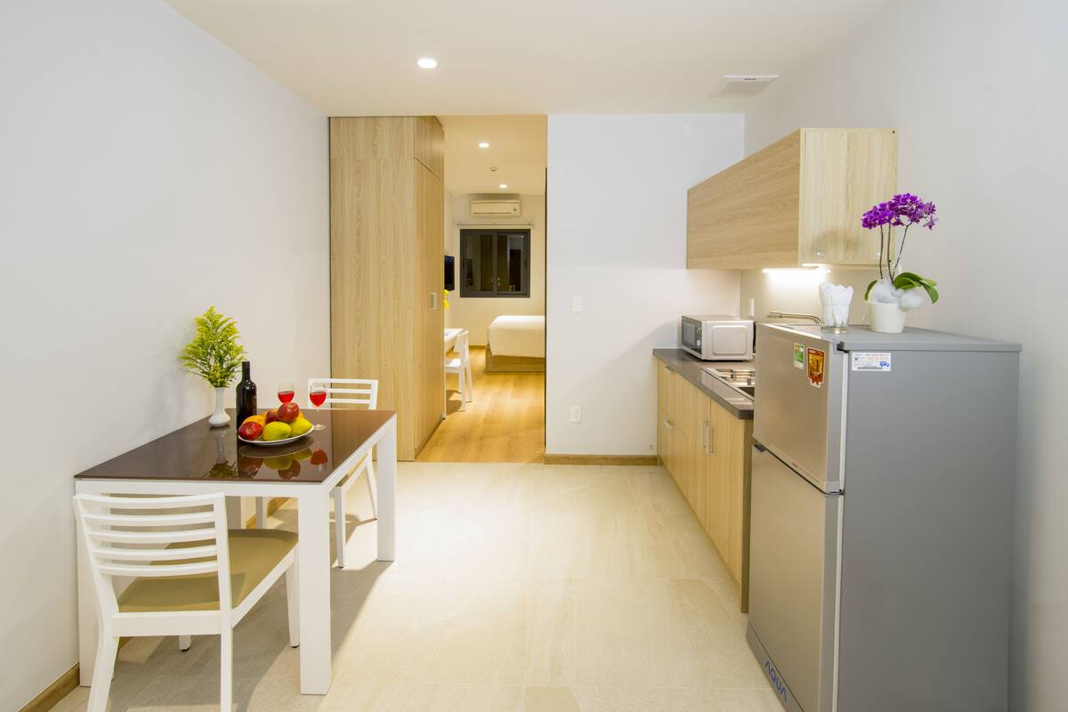 A compact kitchen with a refrigerator, microwave, and wooden cabinets. There is a dining table with chairs and a small potted plant.