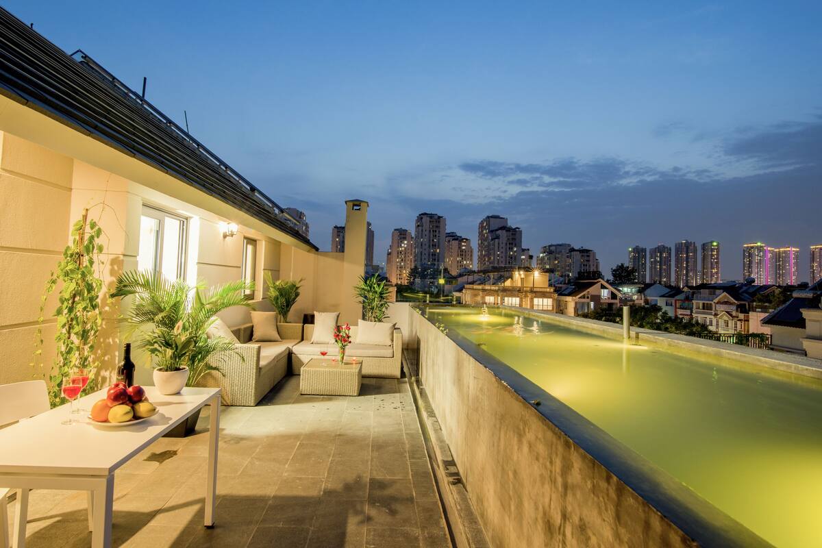 A rooftop terrace with a dining table, seating, and a view of the city skyline at dusk.