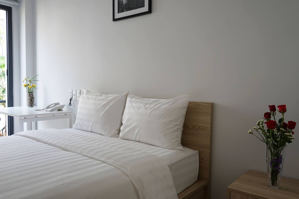 A neatly made bed with white linens, a bedside table with a phone, and a vase with flowers on the nightstand.