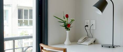 Una habitación con un escritorio blanco, una silla de madera, un florero con flores, una lámpara de escritorio y un teléfono.