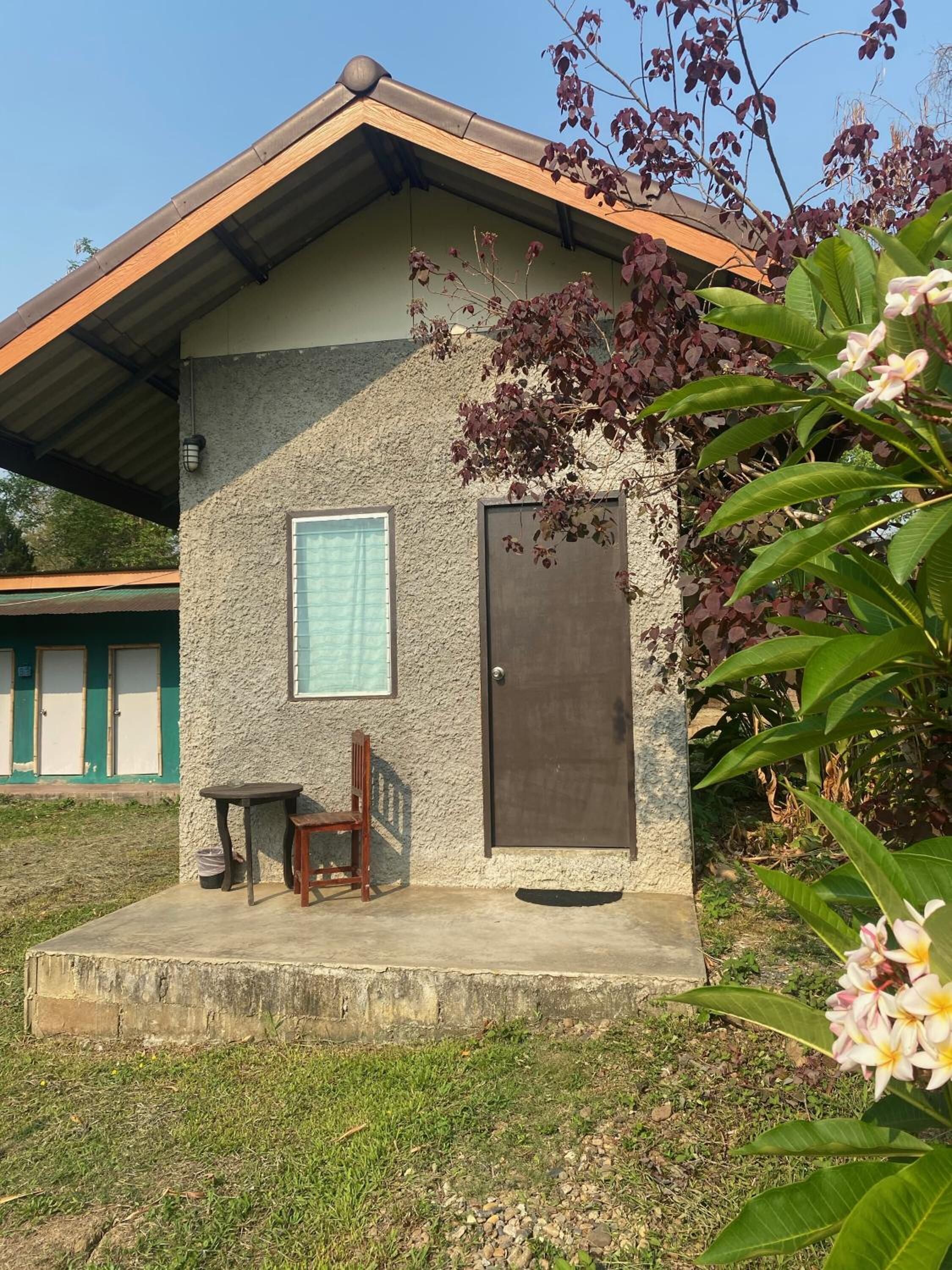 Un pequeño edificio con fachada de concreto, una ventana y una puerta. Hay una pequeña mesa y una silla afuera.