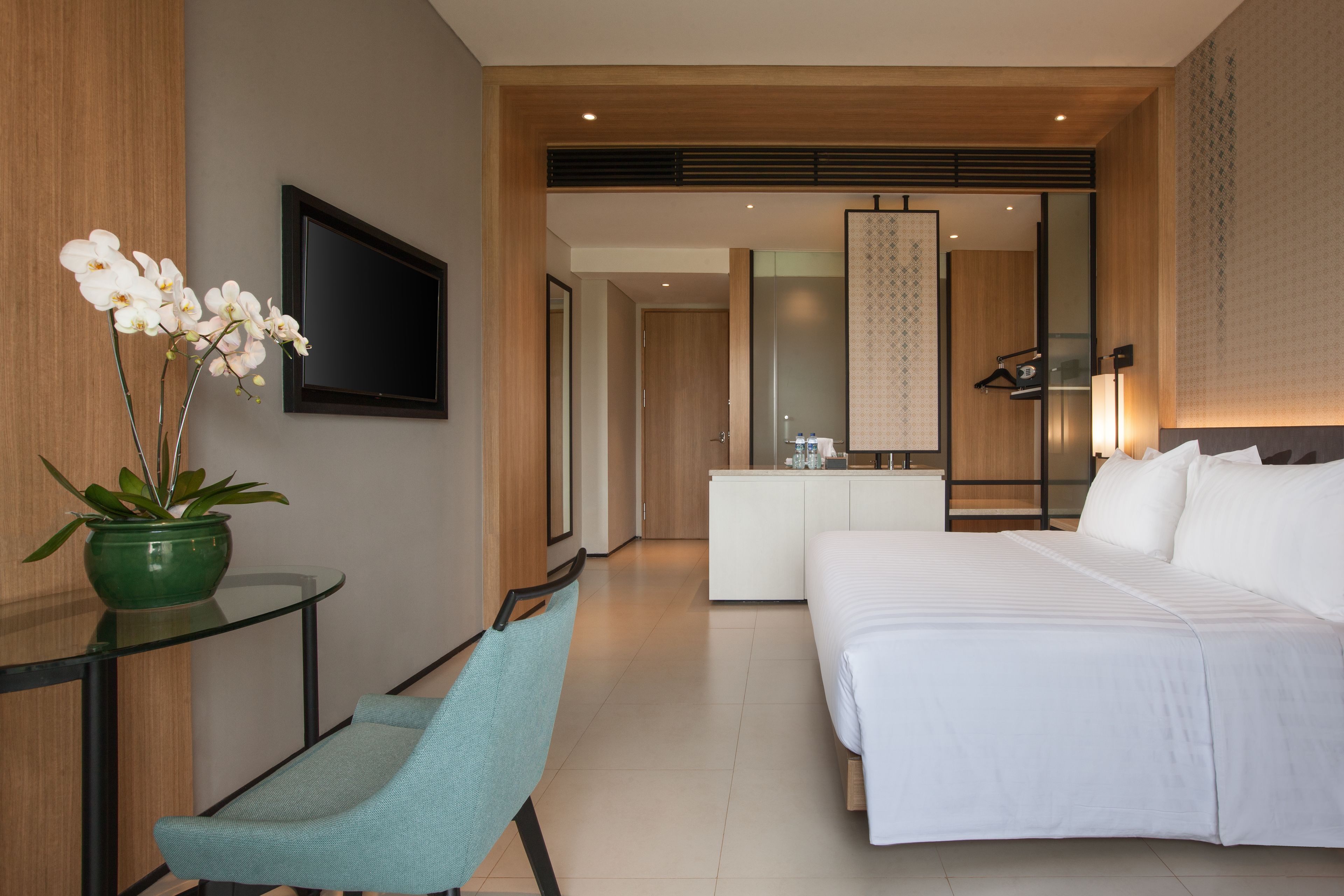 A modern hotel room with a bed, a desk with a chair, a TV, and a bathroom visible through an open door.
