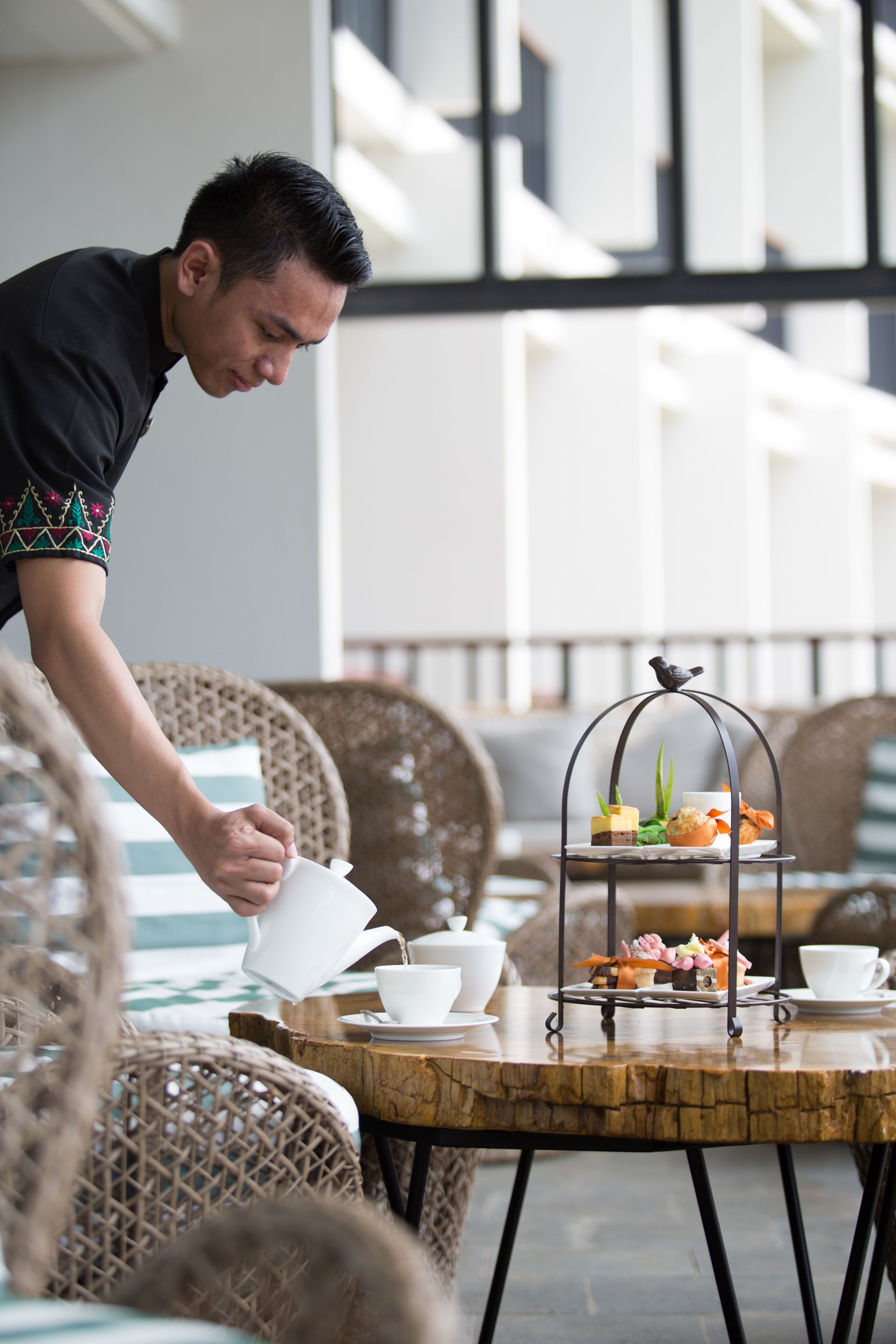 Une personne verse une boisson dans une tasse sur une table où se trouve un présentoir à plusieurs niveaux avec des aliments et une décoration en forme d’oiseau.