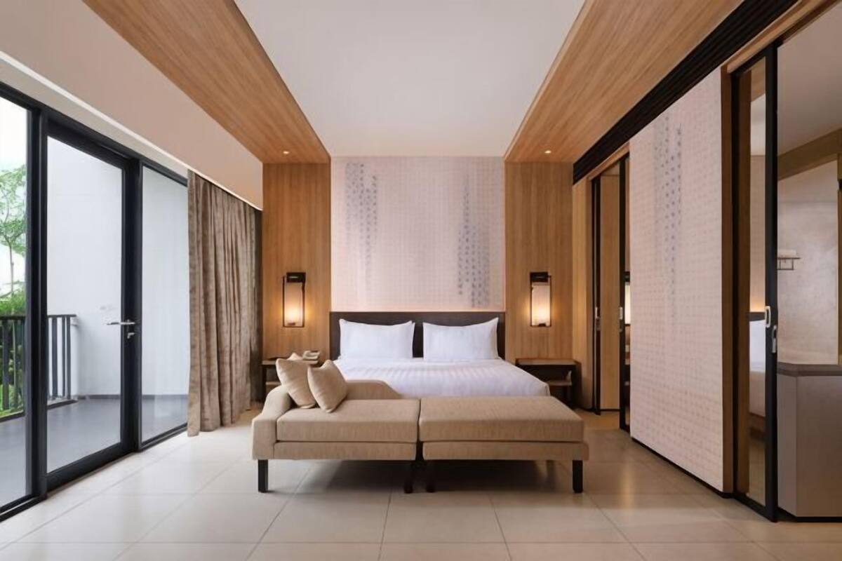 Een moderne hotelkamer met een groot bed, een bank, schuiframen naar een balkon en uitzicht op groen.