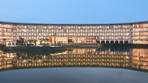 Un grand hôtel à plusieurs étages, avec une piscine dont la surface réfléchit la lumière, située au premier plan.
