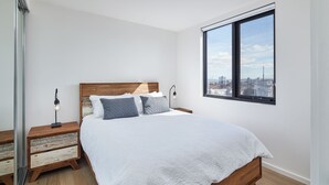 Ein Schlafzimmer mit einem hölzernen Bett, weißer Bettwäsche, einem Nachttisch mit Lampe und einem großen Fenster mit Blick auf die Stadt.