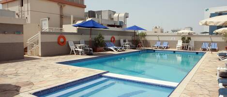 A swimming pool with lounge chairs and umbrellas, surrounded by stone tiles and a building with air conditioning units.