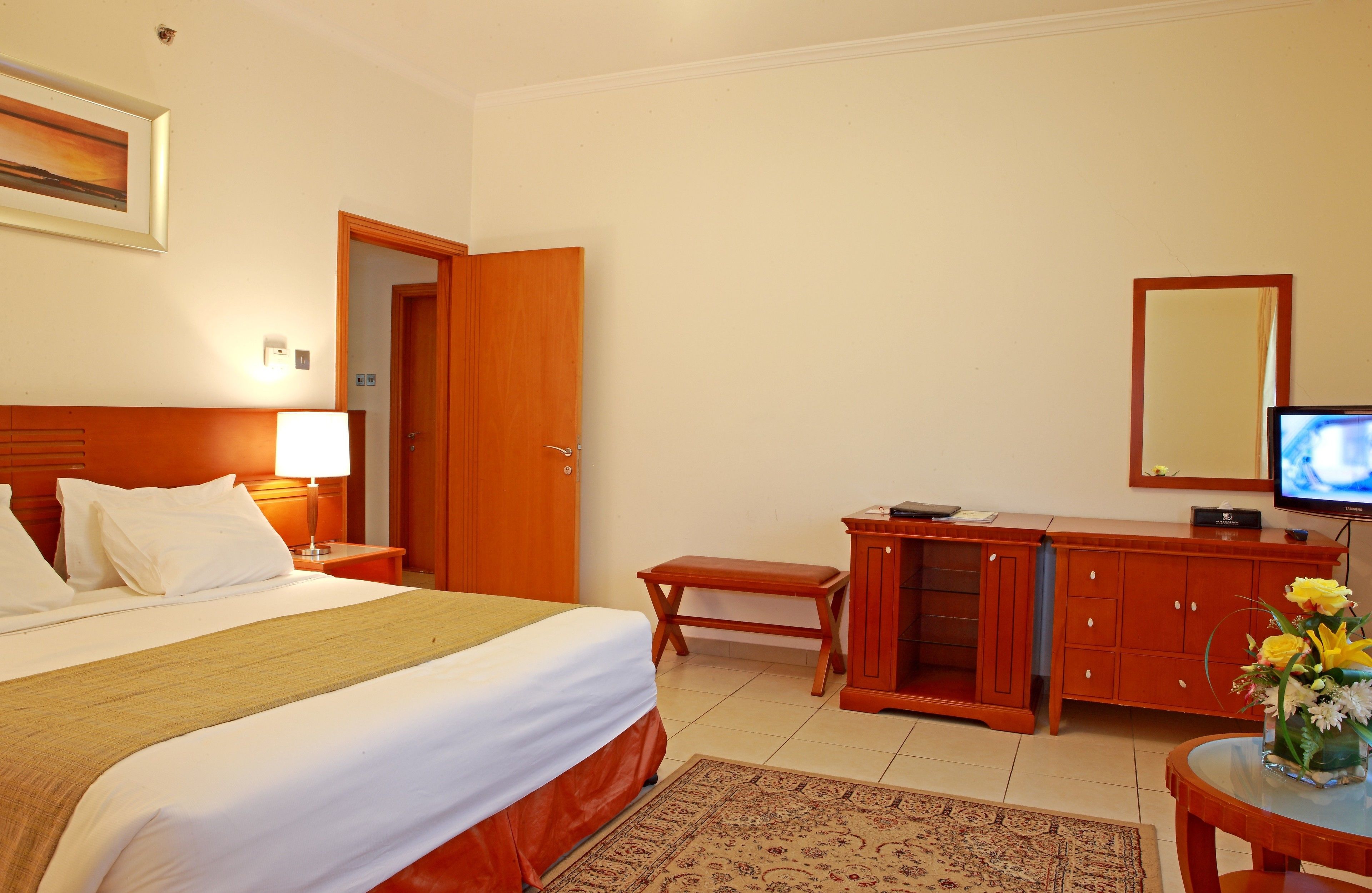 Une chambre d’hôtel avec un lit, une table de chevet, une lampe, un banc en bois, une télévision et une petite table agrémentée de fleurs.