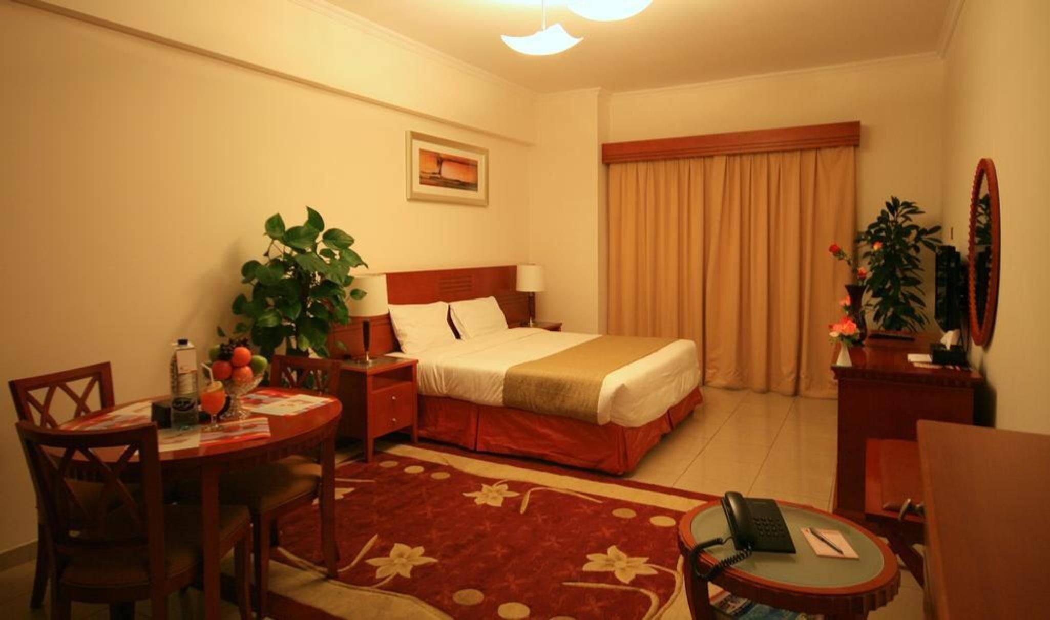 Una habitación de hotel con una cama, un escritorio con una silla, una mesita con una lámpara, un teléfono, un espejo y una planta en maceta.