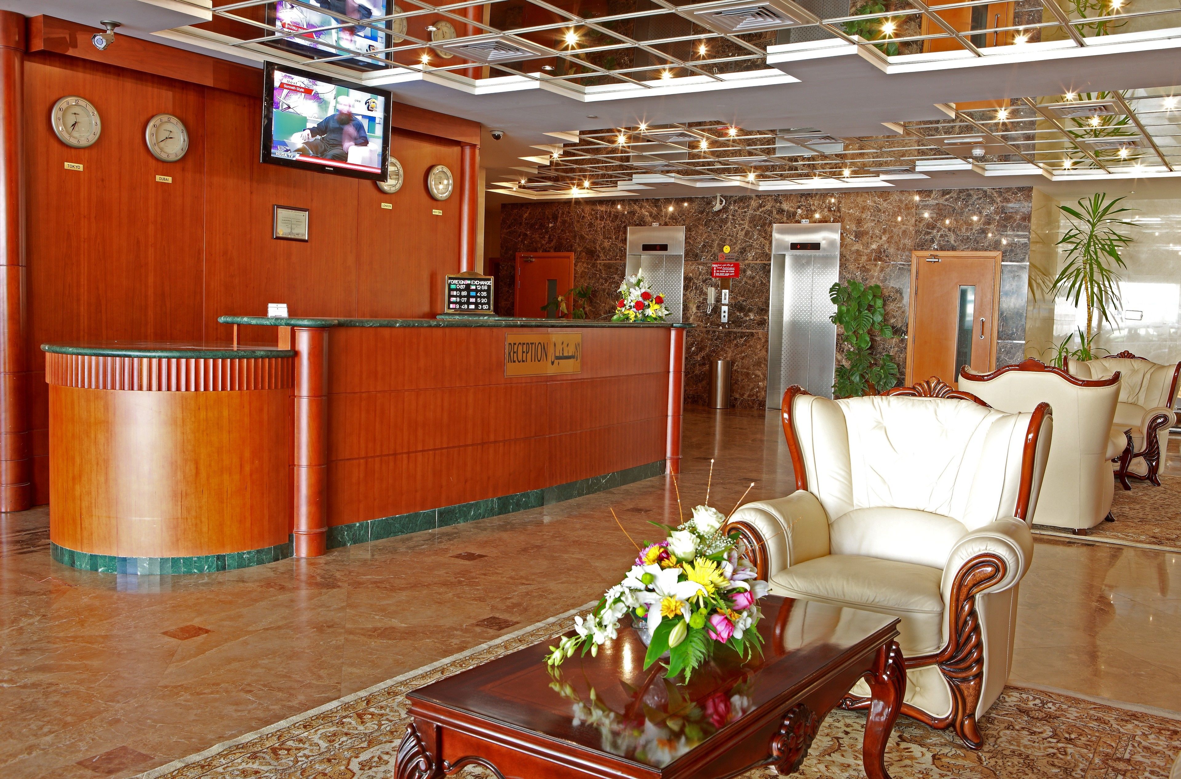 A hotel reception area with a wooden reception desk, a television mounted on the wall, a reception sign, a floral arrangement on a table, and a seating area with a sofa and armchairs.