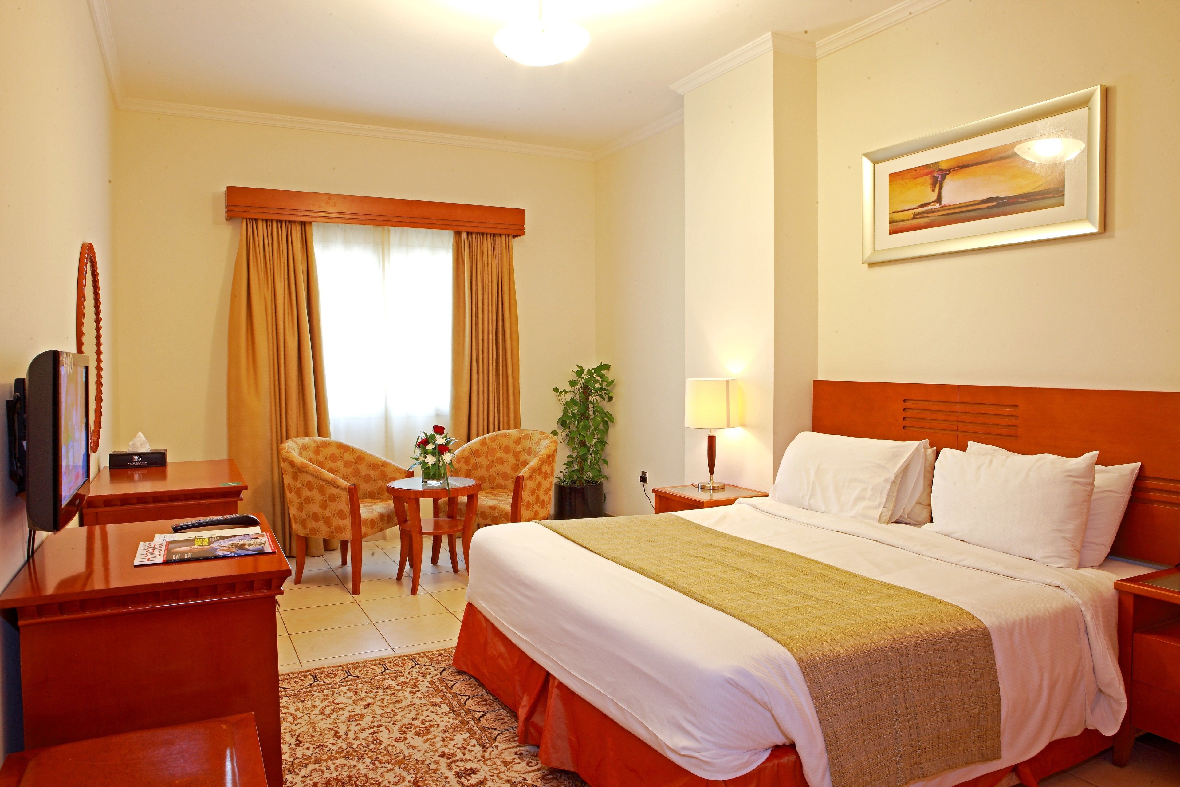 Ein Hotelzimmer mit einem Bett, zwei Sesseln, einem kleinen Tisch mit Blumenvase, einem Schreibtisch mit Fernseher und Fernbedienungen sowie einem Gemälde an der Wand.