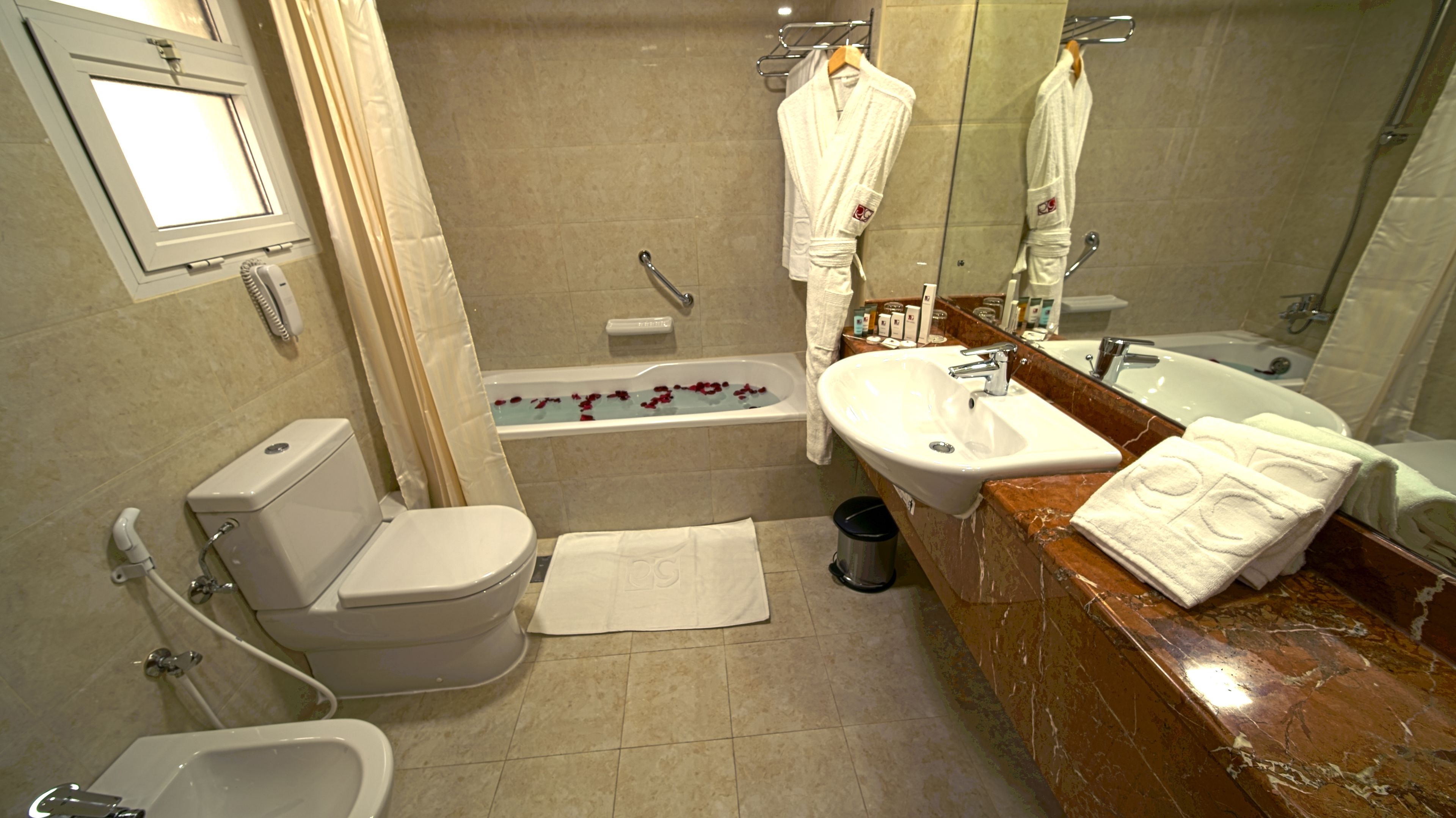 A modern bathroom with a toilet, bidet, bathtub with rose petals, a sink, and a large mirror.
