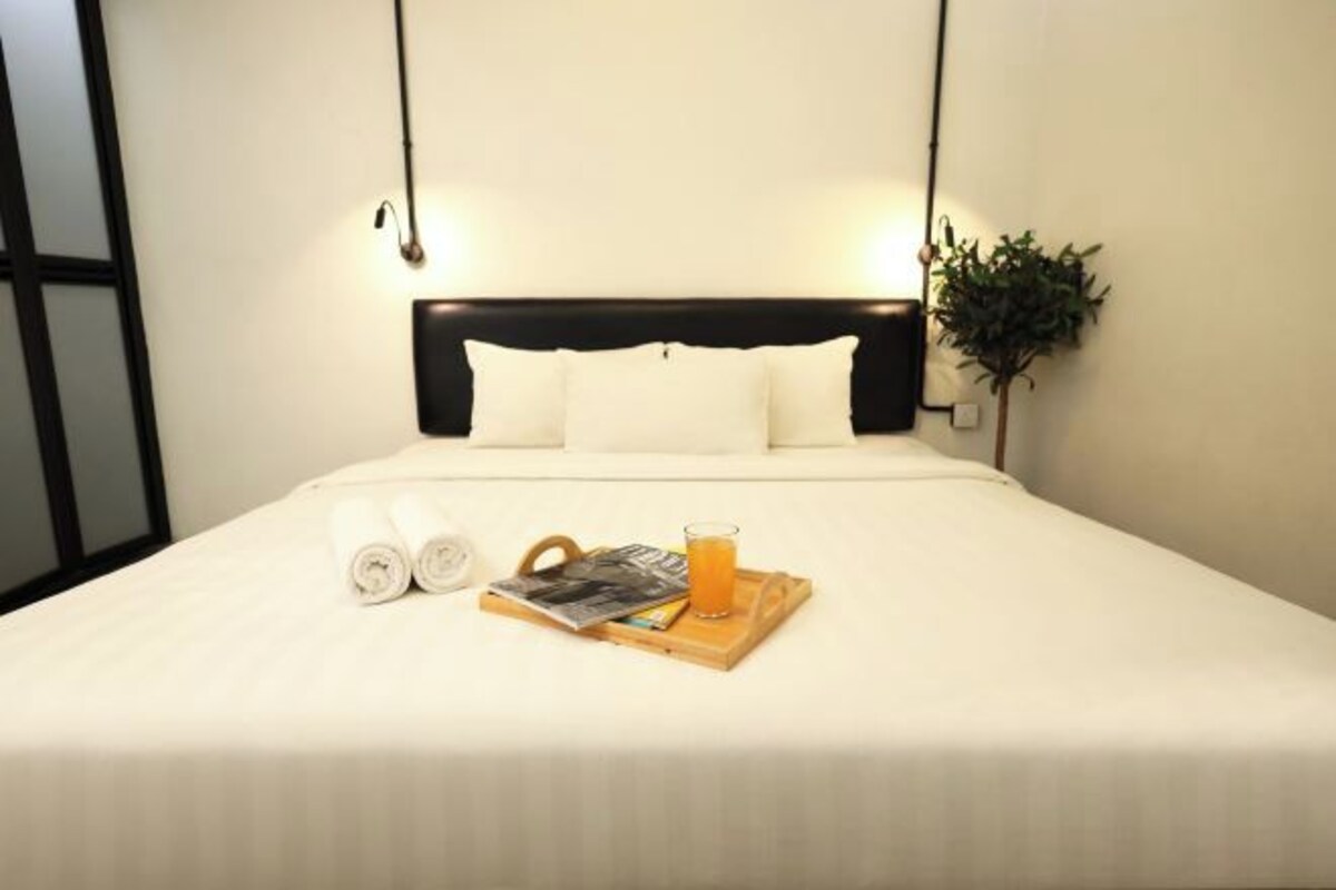 A neatly made bed with white linens, a wooden tray with a magazine and a glass of orange juice, and a potted plant beside the bed.