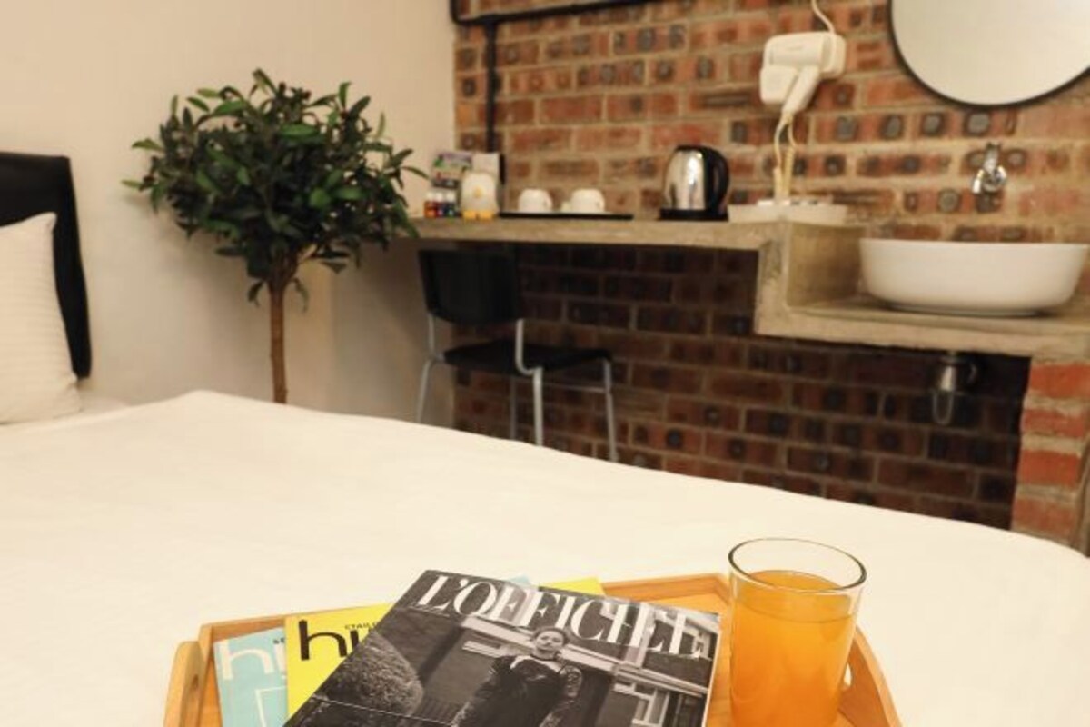 A hotel room with a bed, a bedside table with a tray holding a magazine, a glass of orange juice, and a brick wall with a sink and mirror.