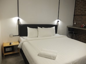 A neatly made bed with white linens and pillows, a small wooden nightstand with a remote control, and a brick wall in the background.