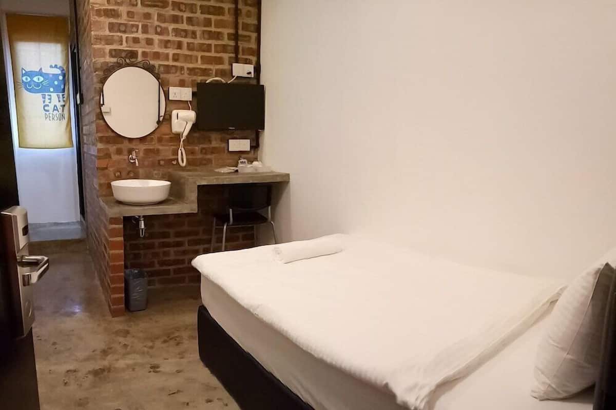 Ein kleines Zimmer mit einem Einzelbett, einer Ziegelwand, einem runden Spiegel, einem weißen Waschbecken und einem Telefon.