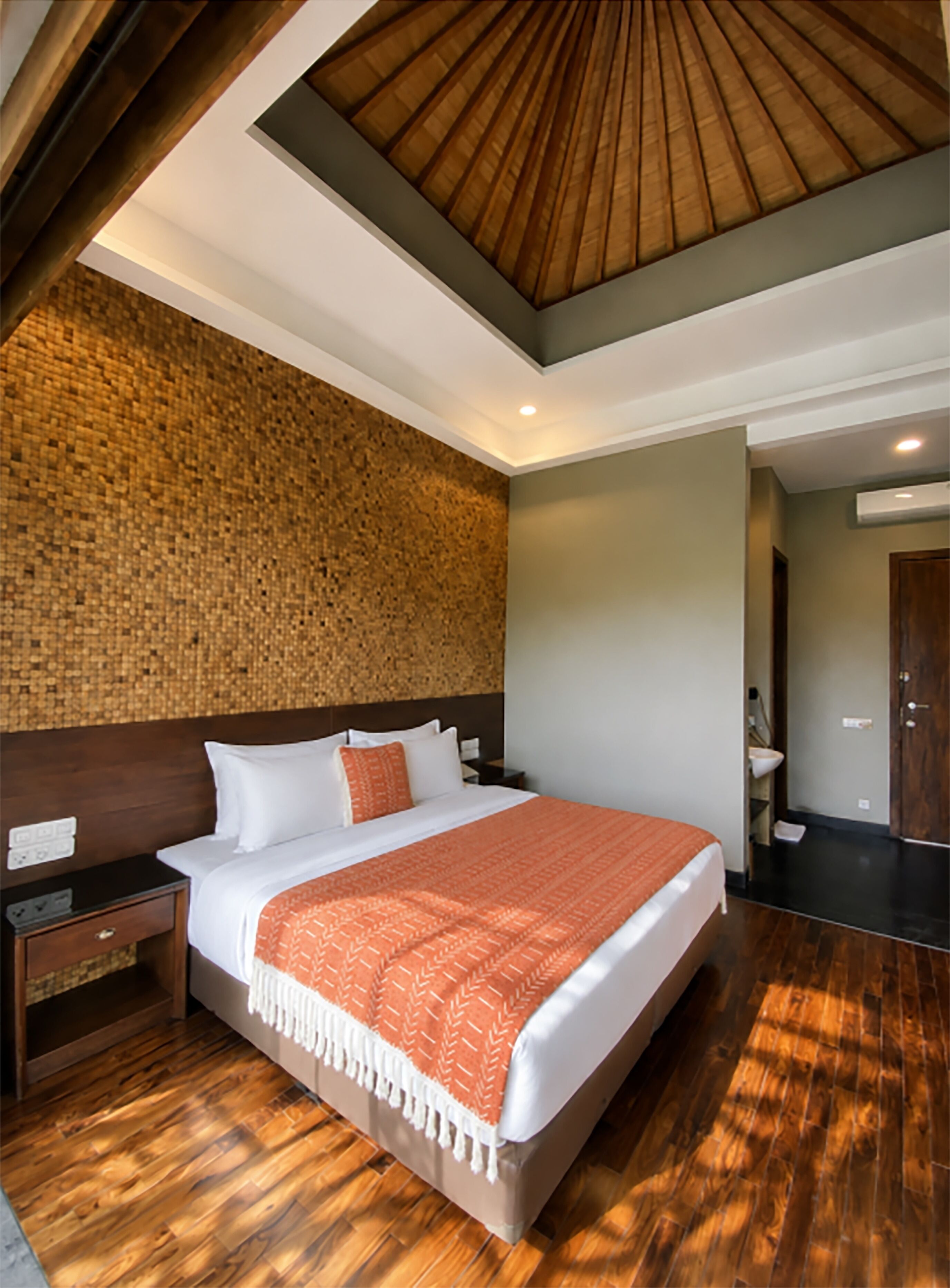 A hotel room with a large bed, wooden flooring, a textured wall, and a modern ceiling design.