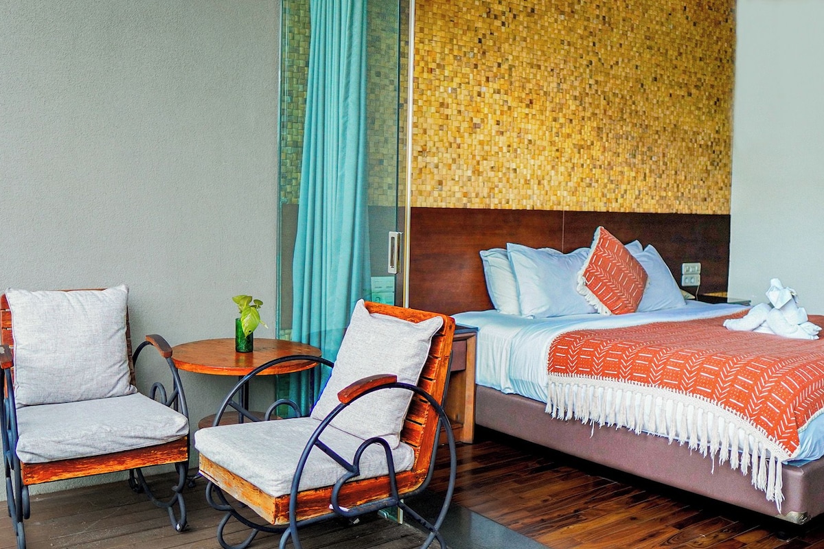 A hotel room with a bed, two chairs, a small table with a plant, and a view of a mosaic wall.