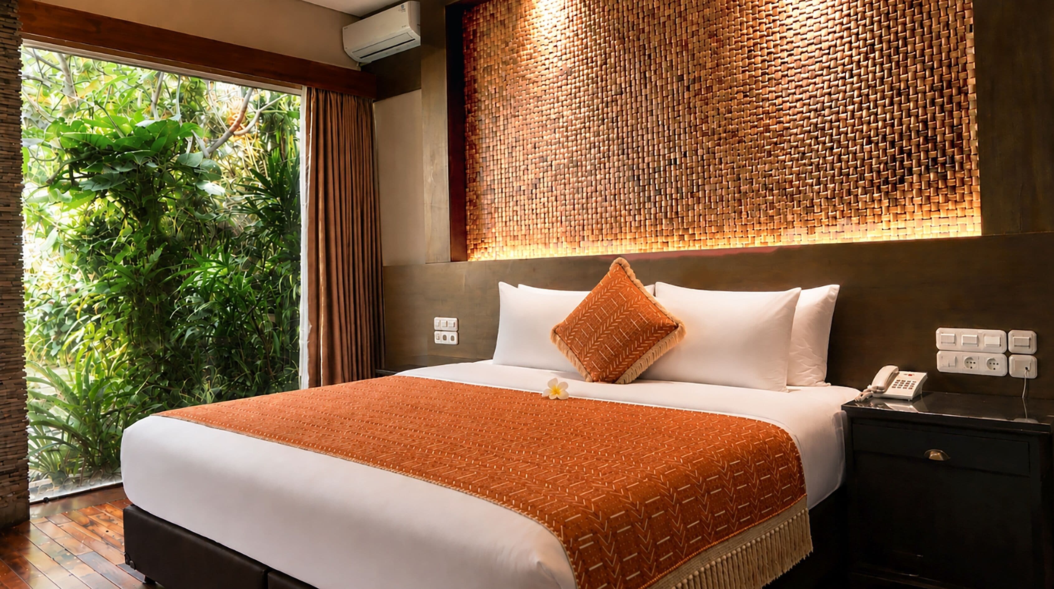 A hotel room with a large bed, a nightstand with a phone, and a view of greenery outside.
