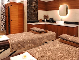 A hotel room with two beds, a wooden wardrobe, a stone accent wall, a sink, and a mirror.