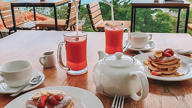 Una mesa de desayuno con tortitas, un jarro de jugo y una tetera, con vistas a un paisaje marino.