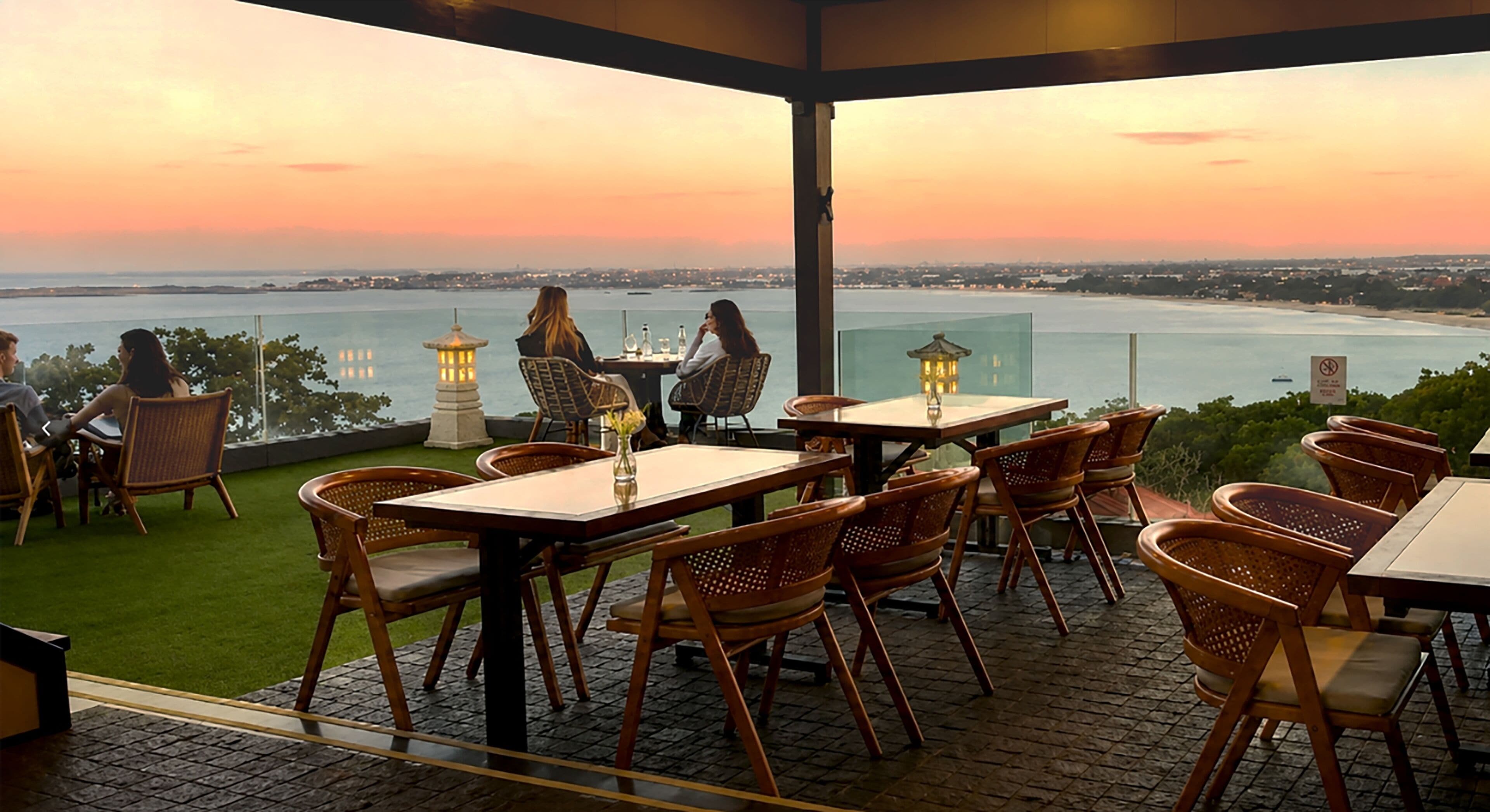 An outdoor seating area with tables and chairs, overlooking a body of water and a distant cityscape.