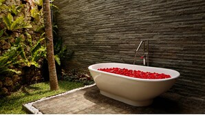 Una bañera independiente llena de pétalos de rosa roja, frente a una pared de piedra con textura, con abundante follaje y una palmera cerca.