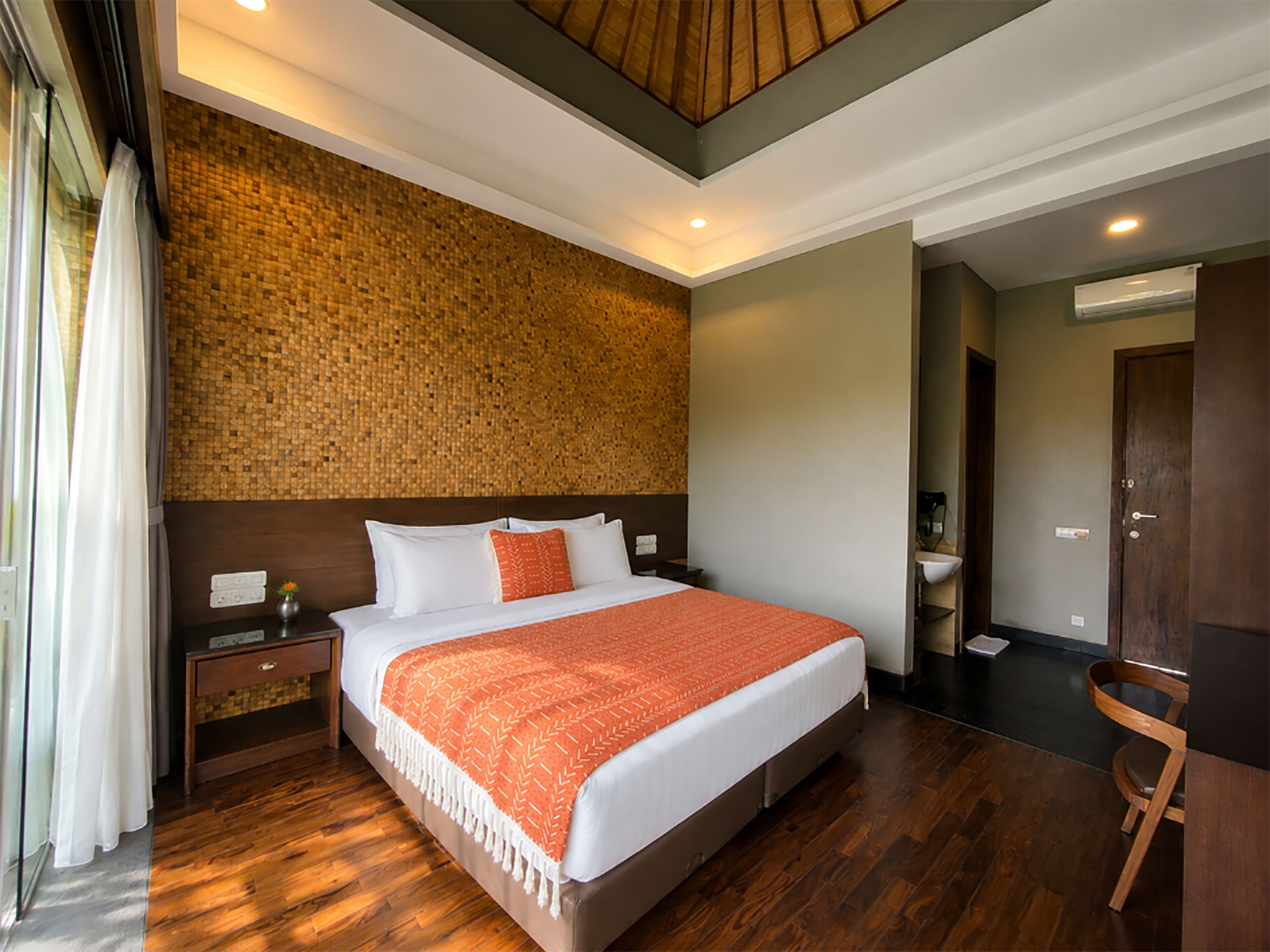 A hotel room with a large bed, wooden flooring, a wall with a textured pattern, a wooden headboard, and a small bedside table with a lamp.