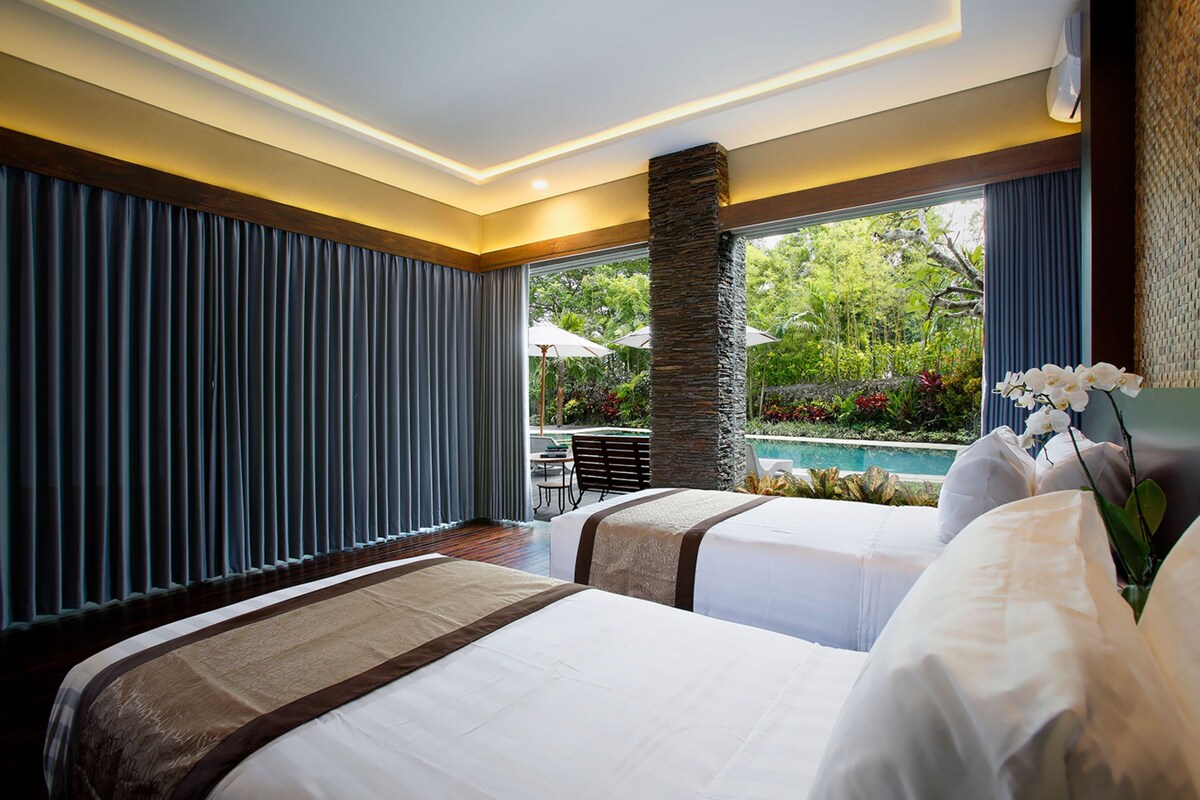 A hotel room with two beds, a stone pillar, a view of a pool, and a dining area with umbrellas.