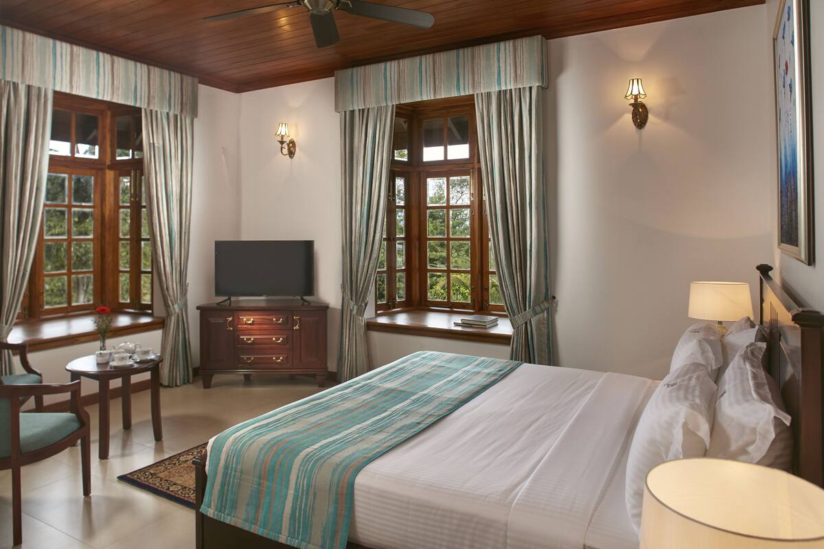 A hotel room with a bed, a TV, a desk, and two windows with curtains.