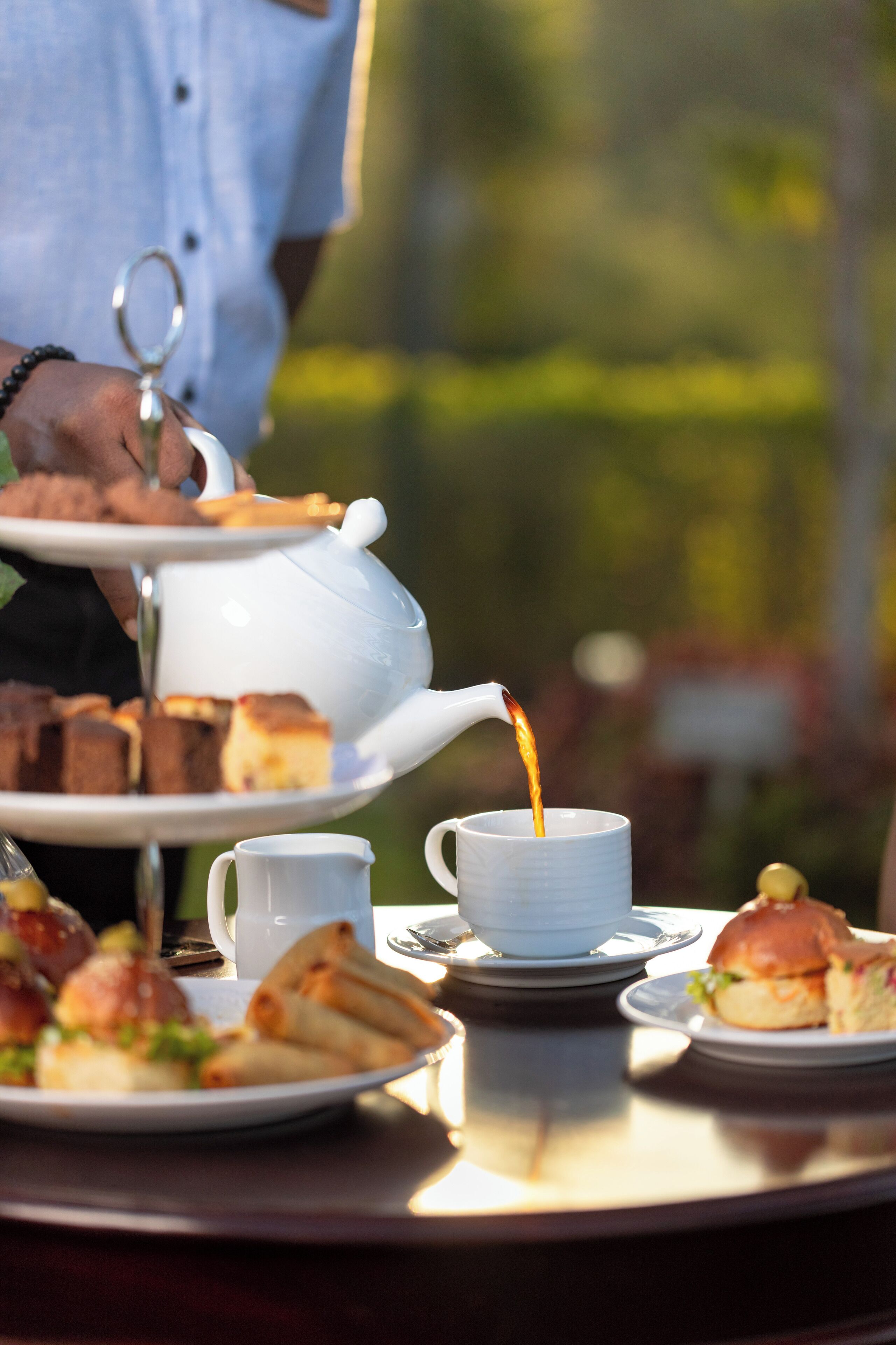 A table with a teapot pouring tea into a cup, a tray with pastries, and sandwiches.