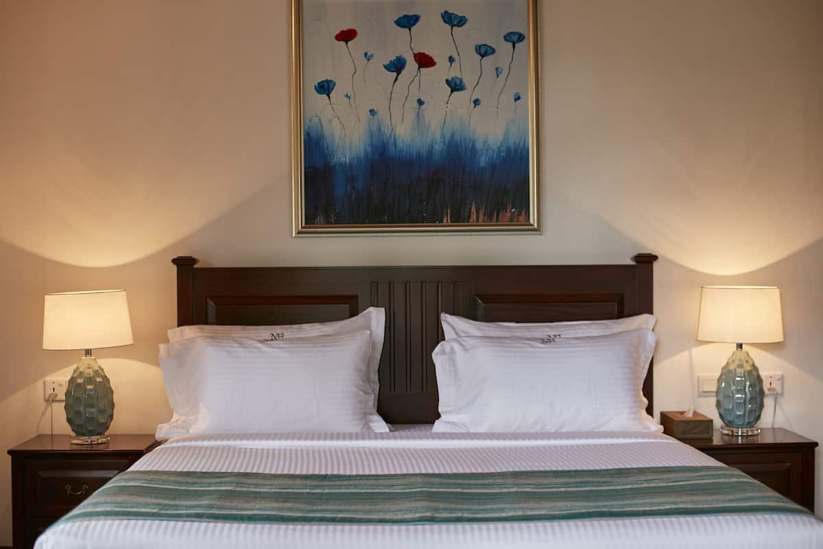 A neatly made bed with white linens, two bedside tables with lamps, and a painting of blue flowers on the wall.