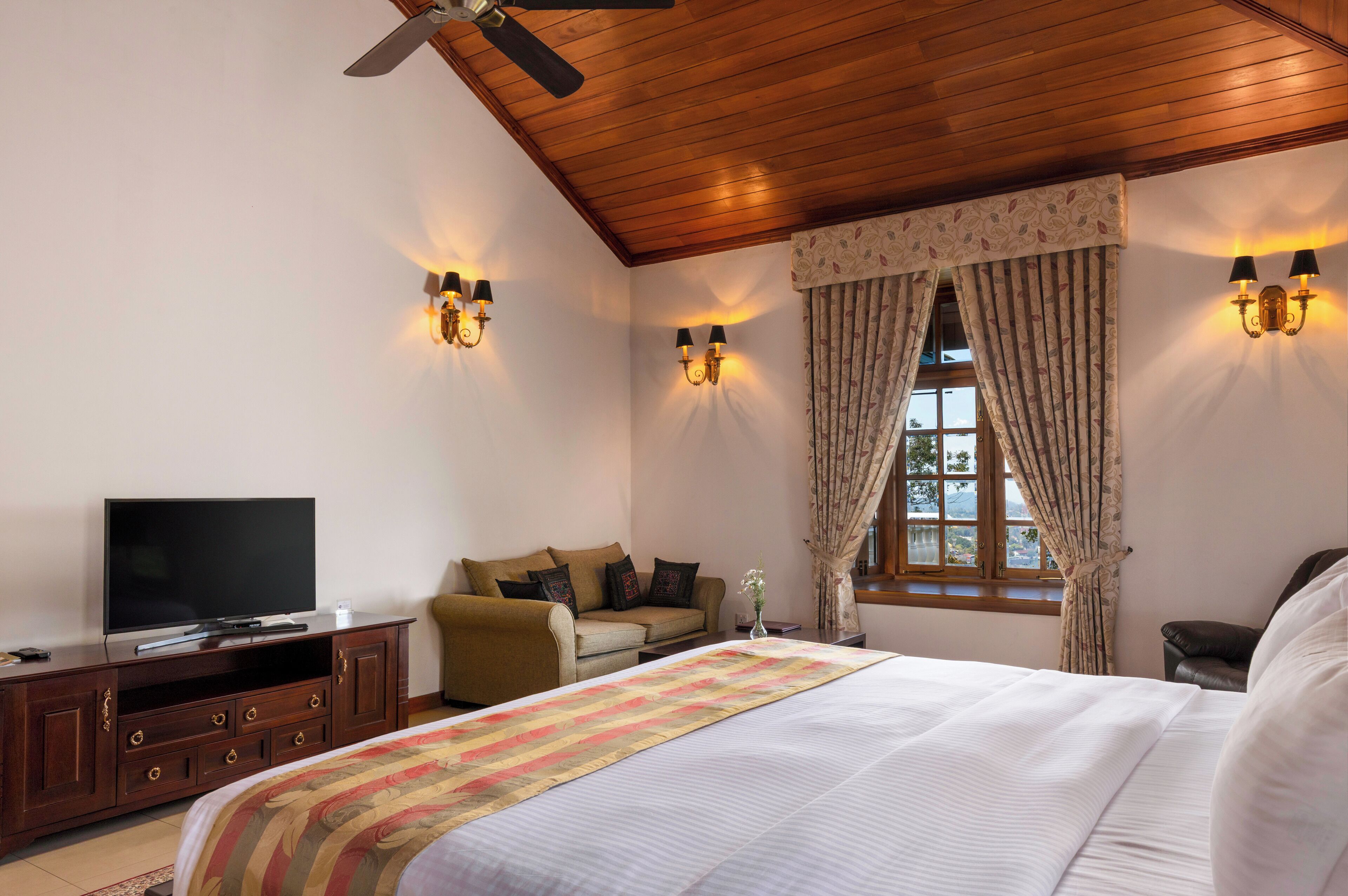 A bedroom with a bed, a television on a wooden cabinet, a ceiling fan, and two wall-mounted lamps.