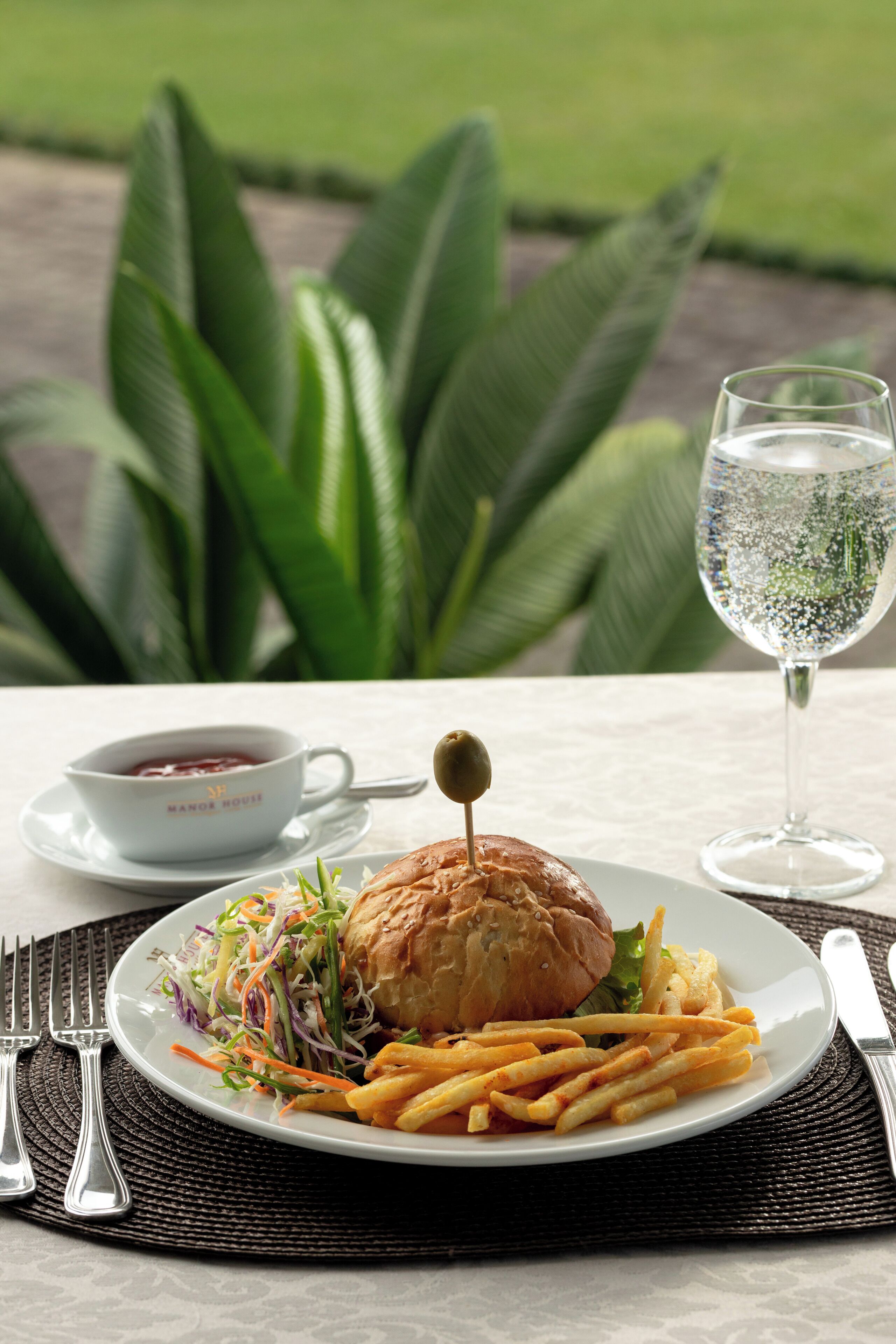 Une assiette contenant un sandwich, des frites et une salade, accompagnée d’un verre d’eau et d’une tasse de thé, est posée sur une table à l’extérieur.