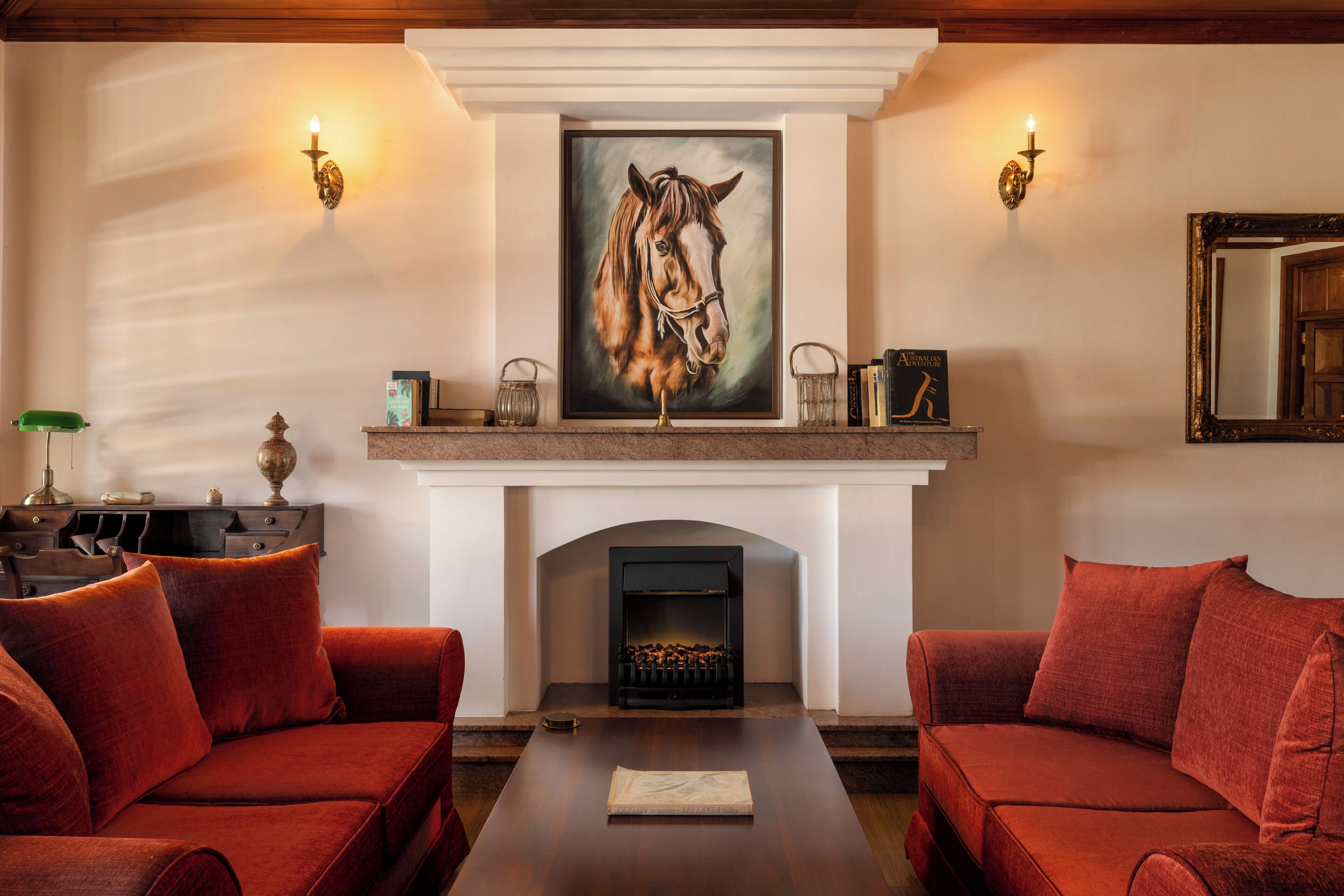 Una habitación con chimenea, dos sofás rojos, una mesa y un cuadro enmarcado de un caballo.