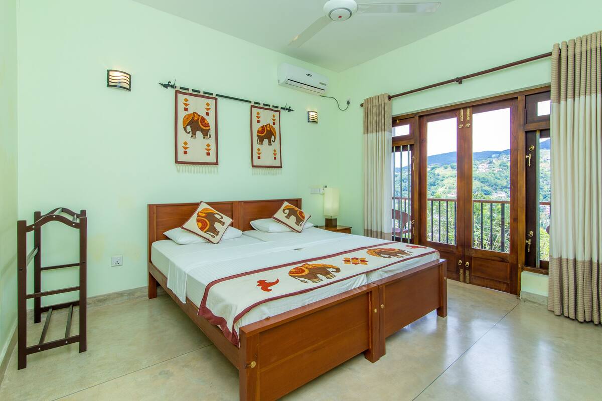 A bedroom with a large bed, a wooden ladder, a ceiling fan, and a view of the outdoors through a door.