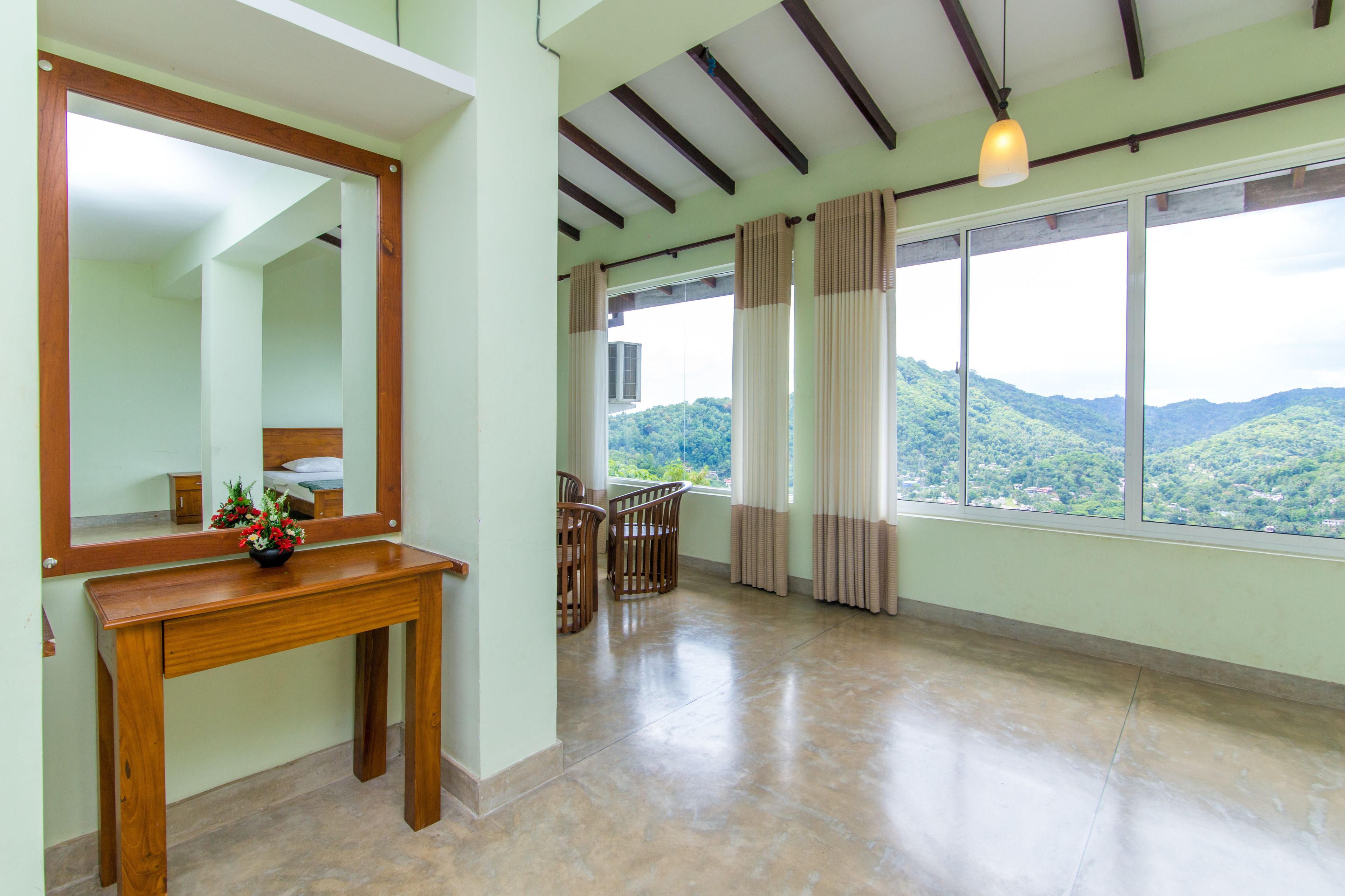 Una habitación amplia con un gran ventanal que ofrece vistas a colinas verdes, una mesa de madera con un florero y un espejo en la pared.
