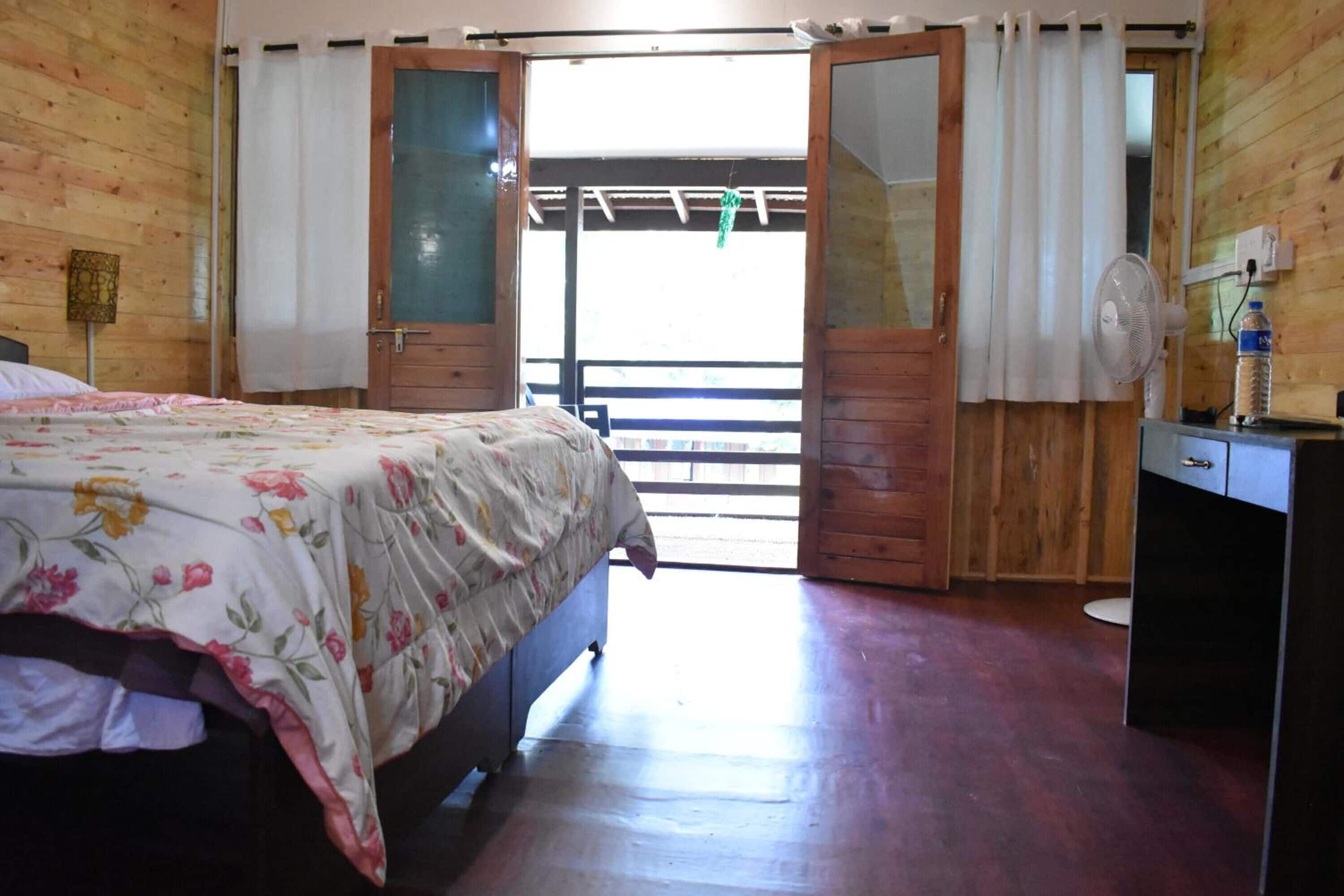 A bedroom with a bed, a wooden wardrobe, a desk with a fan, and a window with curtains.