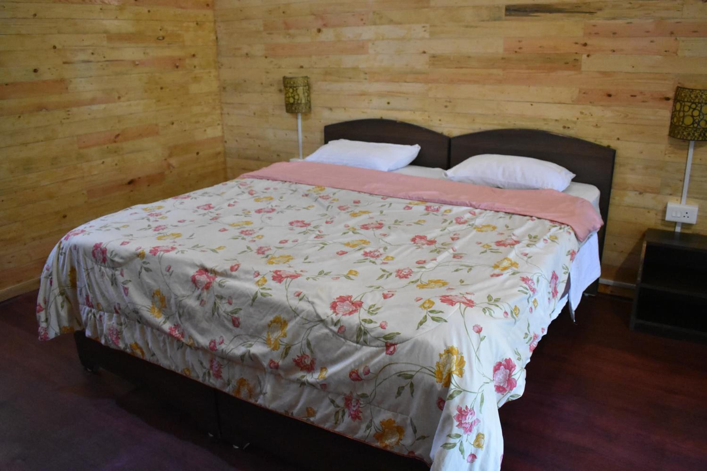A bedroom with a wooden wall, a bed with floral bedding, and a nightstand with a lamp.