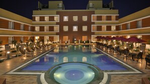 Piscina com espreguiçadeiras e guarda-sóis, rodeada por um edifício com varandas e janelas.