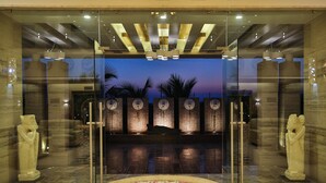 Entrada de hotel moderna com portas de vidro, um arranjo floral decorativo e vista para palmeiras ao entardecer.