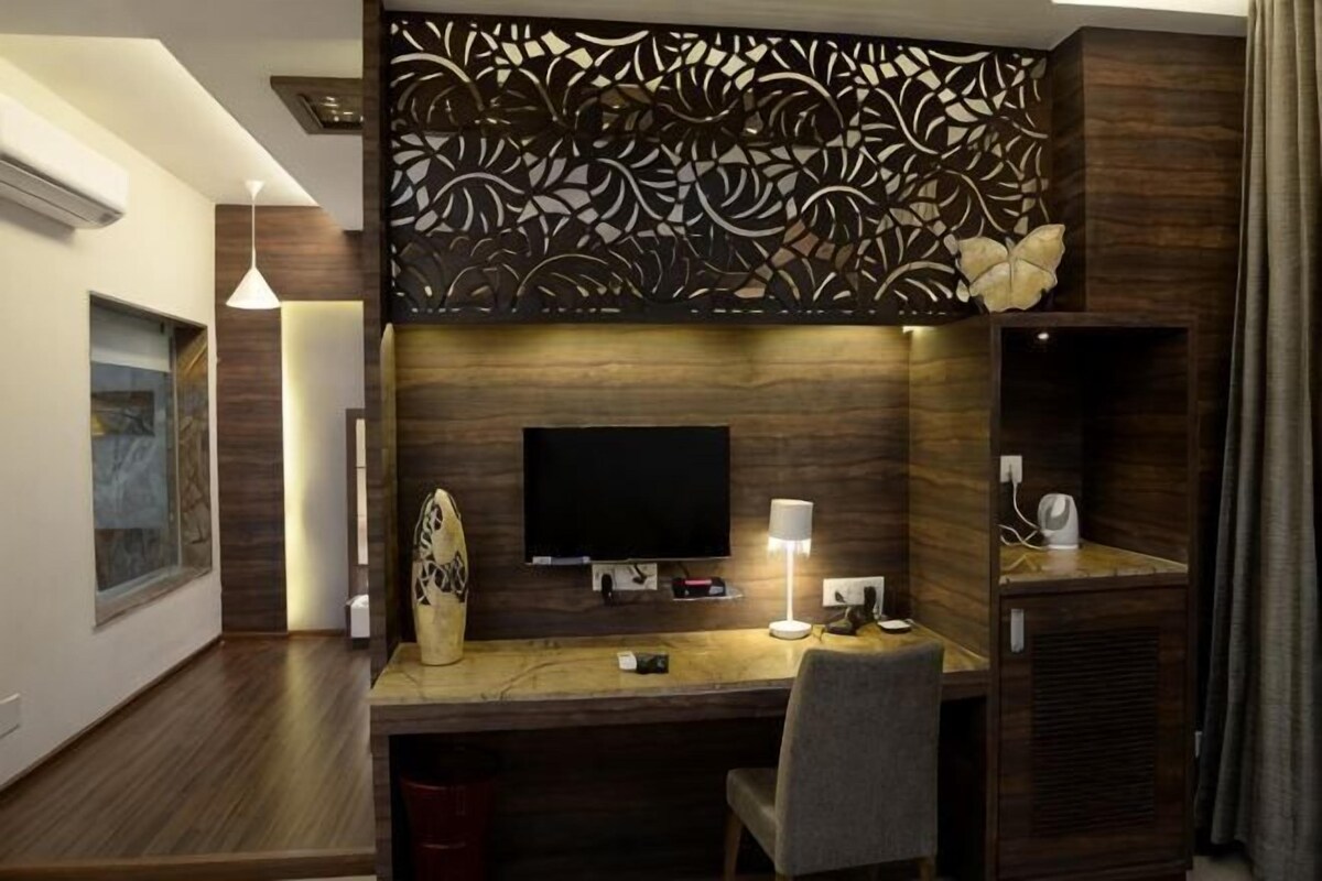 A modern hotel room with a wooden desk, a flat-screen TV, a lamp, and a decorative wall panel.