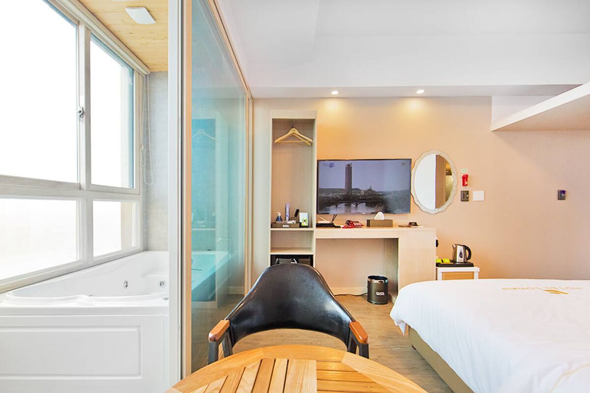 Una habitación de hotel moderna con una cama grande, un escritorio con silla, un televisor y vistas a un paisaje urbano a través de una ventana.