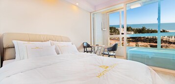 A hotel room with a bed, a small table, two chairs, and a large window offering a beach view.