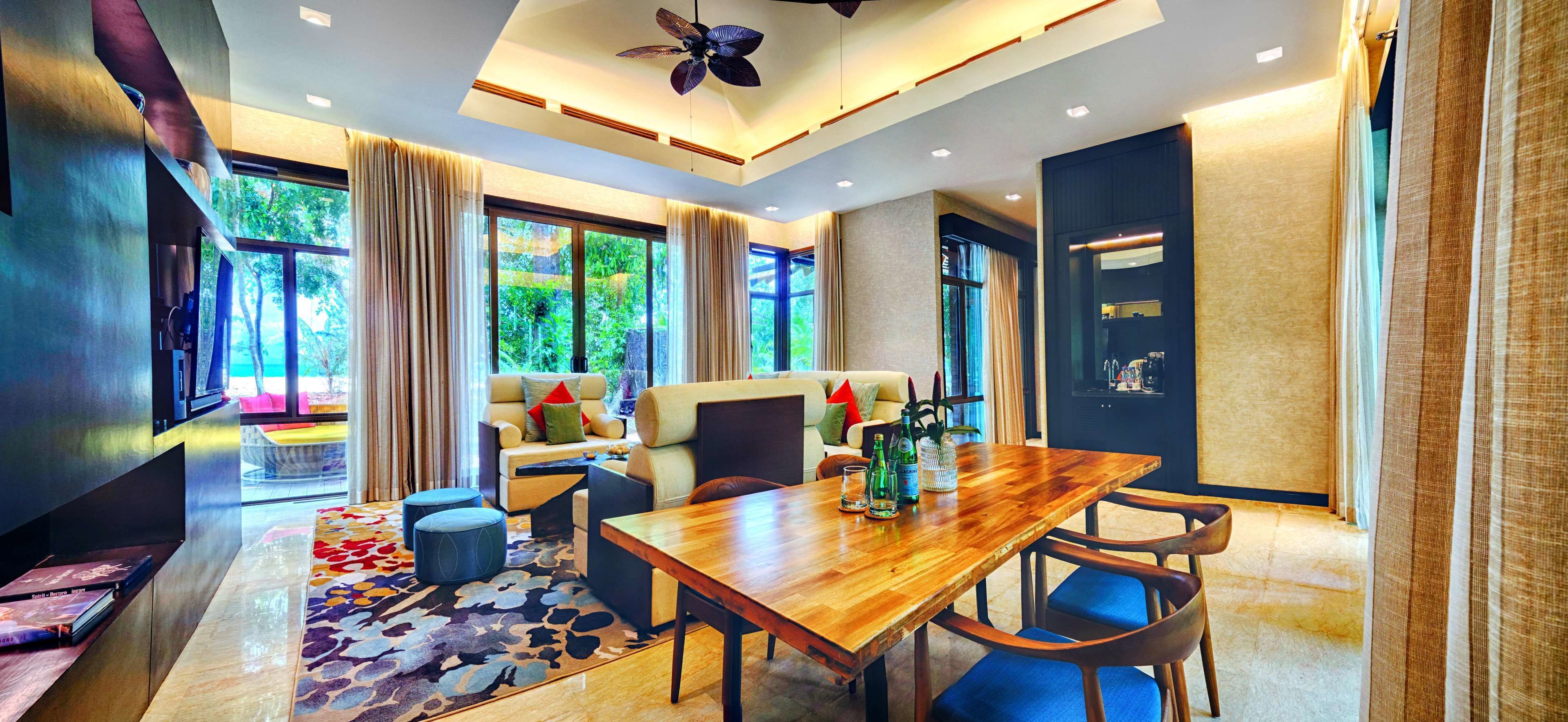 A modern hotel room with a wooden dining table, comfortable seating, large windows with curtains, and a ceiling fan.