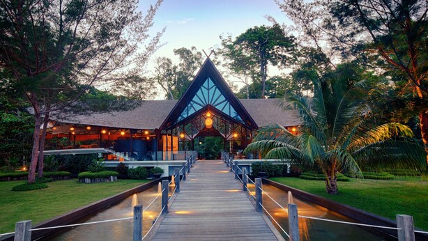 A wooden pathway leading to a building with a triangular roof and large windows, surrounded by lush greenery and palm trees. - Borneo Eagle Resort (Kota Kinabalu)