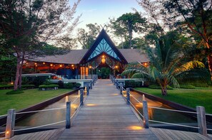 A wooden pathway leading to a building with a triangular roof and large windows, surrounded by lush greenery and palm trees. - Borneo Eagle Resort (Kota Kinabalu)